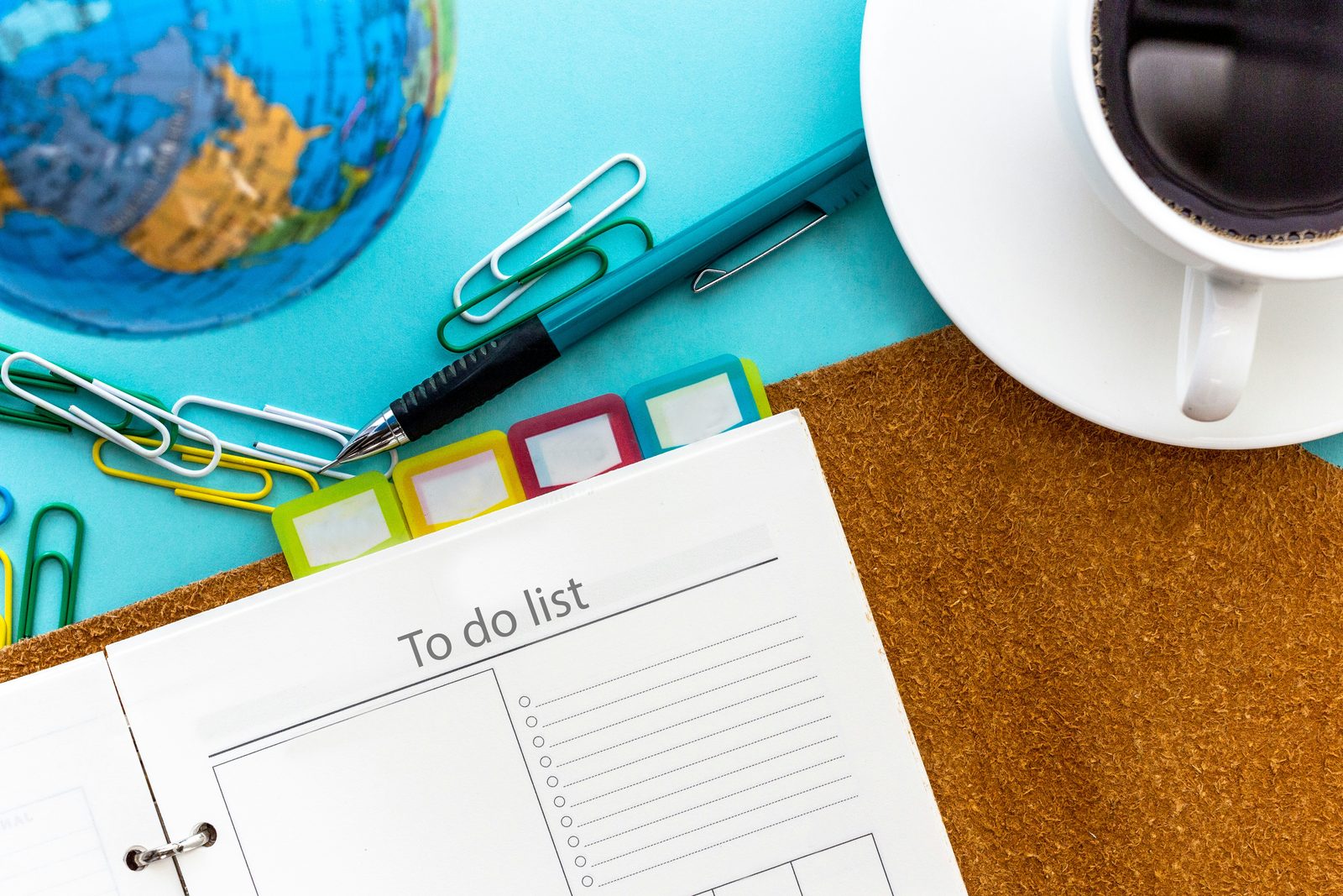 A to-do list notepad on a desk beside a pen, colored paper clips, tabbed dividers, a globe and a cup of coffee.