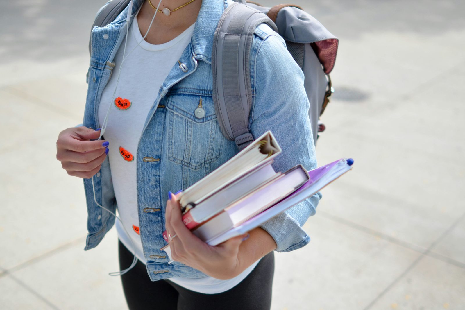 A student outside school carrying a stack of books and notebooks, wearing a backpack and a denim jacket.